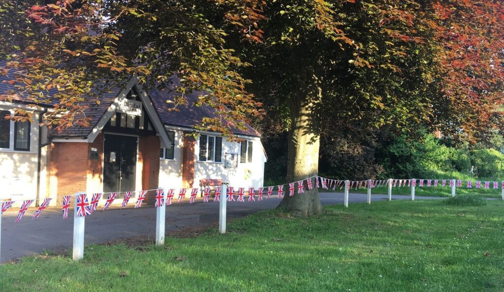 Callow End Village Hall, a white-rendered hall with a timbered gable porch, under a copper beech, with Union Jack bunting strung along the white fence posts in front