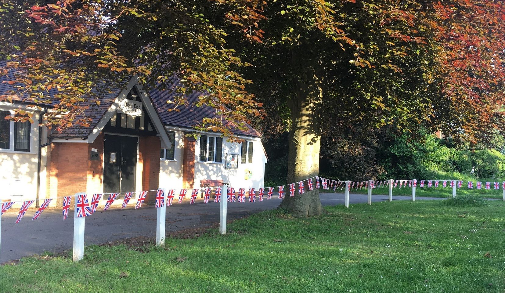 Callow End Village Hall, a white-rendered hall with a timbered gable porch, under a copper beech, with Union Jack bunting strung along the white fence posts in front